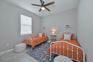 A bedroom with two beds, a ceiling fan, a window with blinds, a bedside table with a lamp, and a decorative wall hanging. - New - Lux - Waterfront - Hot Tub - Bikes (Cape Coral)