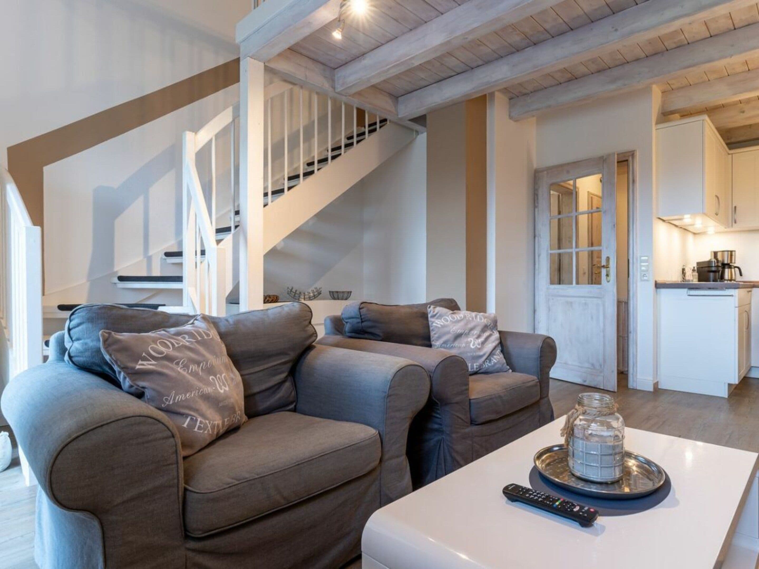 A modern living room with a staircase, a kitchen area, and a dining table with a remote control.