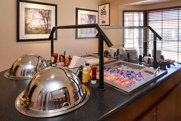 A breakfast buffet area with covered food stations, condiments, and a variety of yogurt cups.