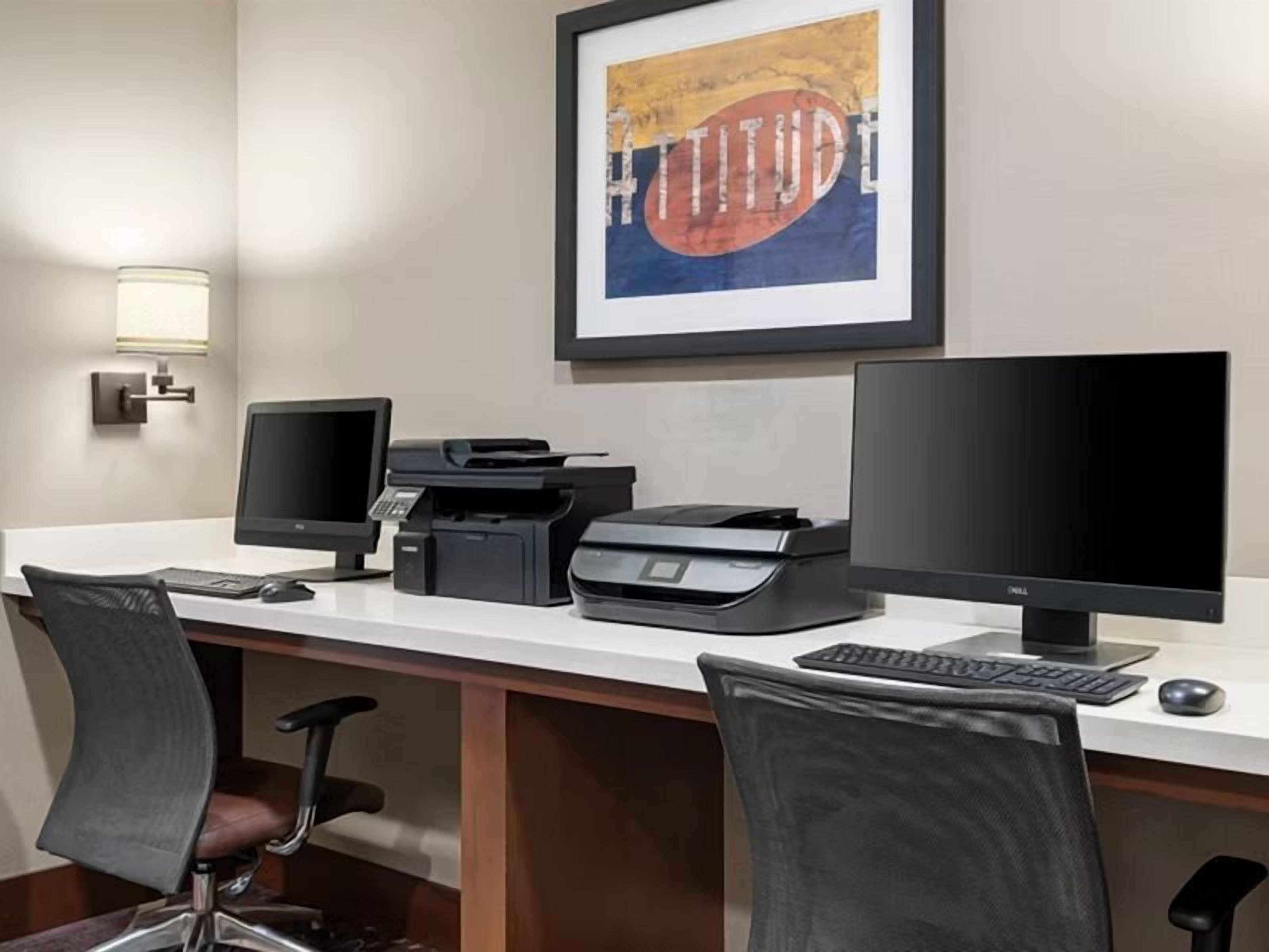 Una oficina de hotel con dos monitores de computadora, una impresora, un teclado y una silla.