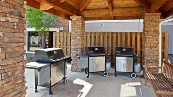 An outdoor patio with two barbecue grills, a stone pillar, and a wooden bench.