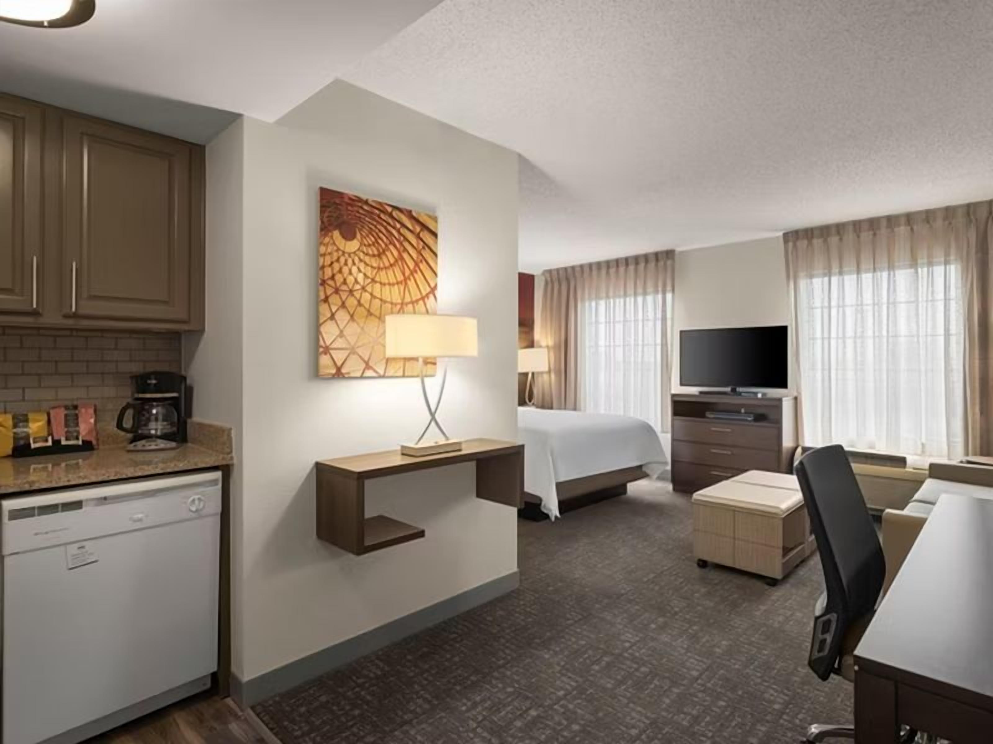 A hotel room with a kitchenette, a bed, a desk, a chair, a TV, and a window with curtains.