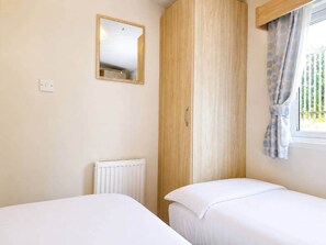 A bedroom with two beds, a wooden wardrobe, a radiator, a mirror, and a window with curtains. - The Retreat Anchorage – 8-Berth Caravan (Rhyl)
