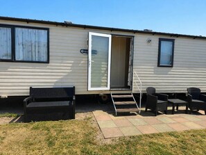 A mobile home with a sliding door, a small outdoor seating area with a bench and chairs, and a paved patio. - The Retreat Prestatyn Beach – 3-Bed (Prestatyn)