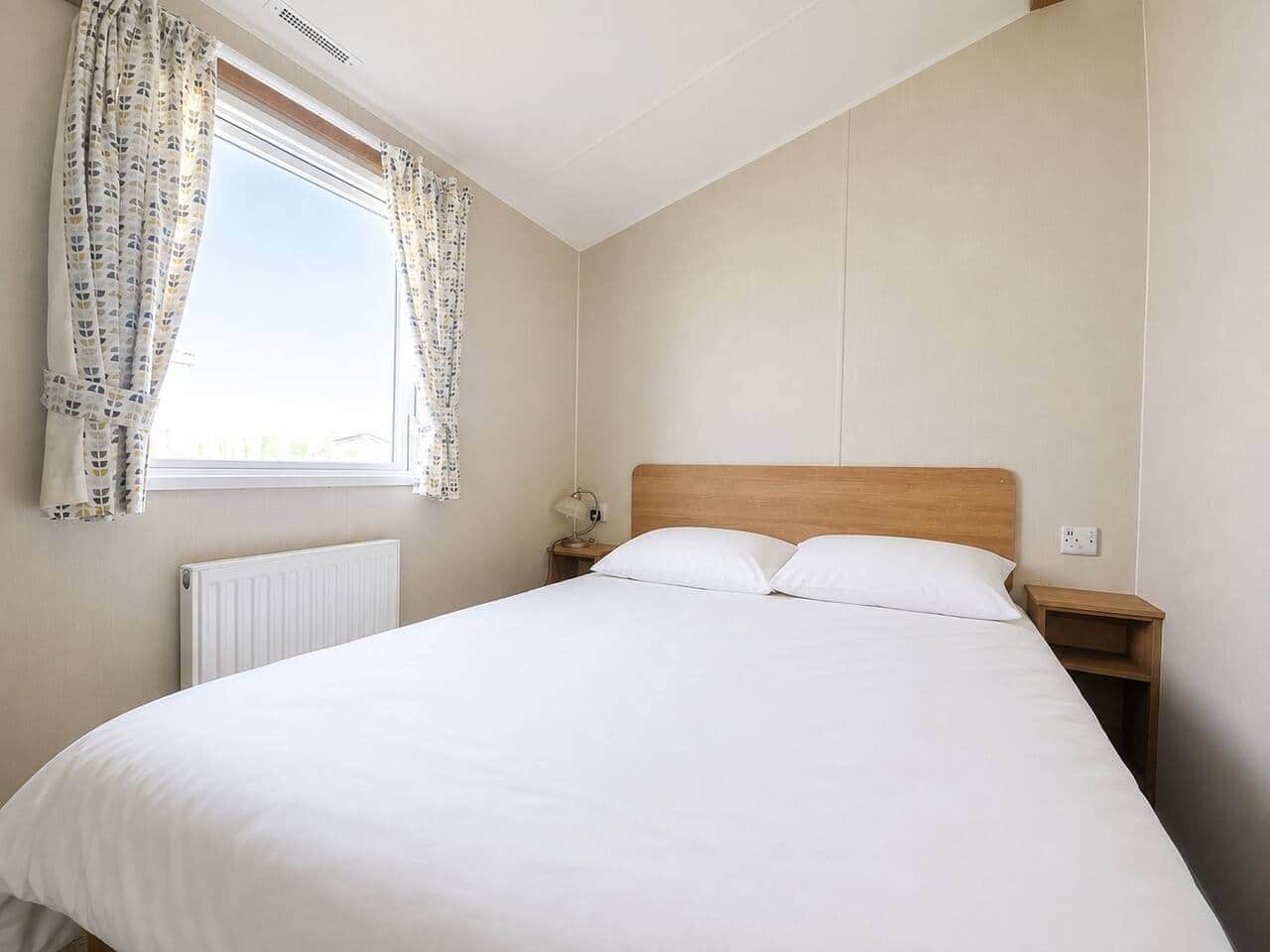 A single-bed room with a wooden headboard, a bedside table with a lamp, and a window with patterned curtains.