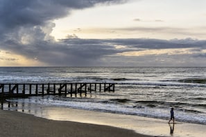 Ein Strand mit einer hölzernen Steg, der sich ins Meer erstreckt, Wellen schlagen an das Ufer und eine Person geht am Strand entlang.