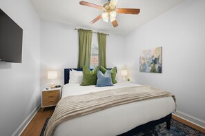 A hotel room with a bed, nightstands, a ceiling fan, a window with curtains, a painting, and a view of a building outside.