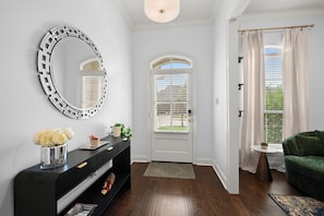 A hallway with a black console, a round mirror, a white door with a window, and a green sofa. - Modern LSU Retreat | 3 Kings Pool Gated Community (Baton Rouge)