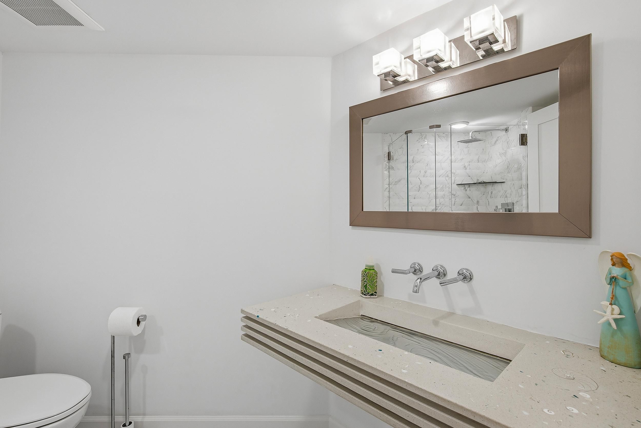 A bathroom with a toilet, a sink with a marble countertop, a shower with a glass door, and a wall-mounted mirror with lights.