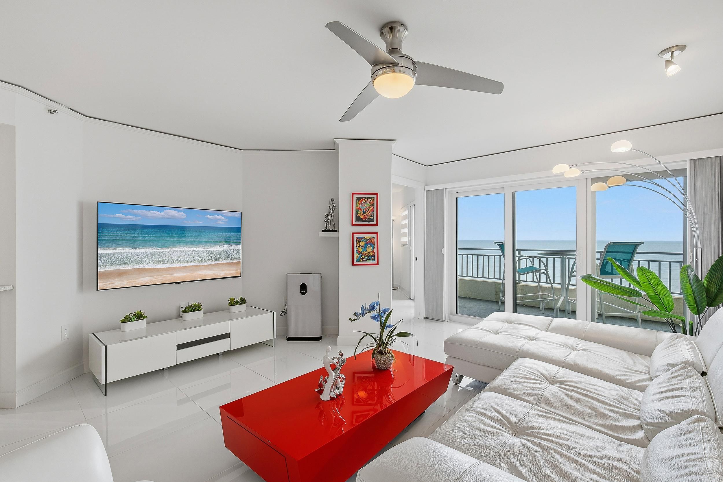 A modern living room with a flat-screen TV, a ceiling fan, and a view of the ocean through large windows.