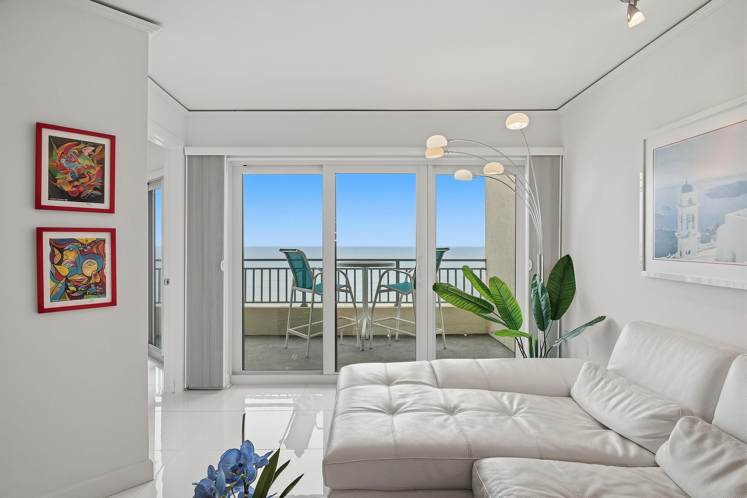 Un salon moderne avec un canapé blanc, un balcon donnant sur la mer et deux œuvres d’art encadrées accrochées au mur.