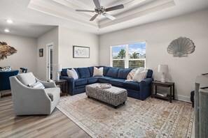 A living room with a sectional sofa, a grey armchair, a patterned ottoman, a ceiling fan, and a view of the outdoors through the windows. - Endless Summer | Beach, Sleeps 14, Game Room (Port Saint Joe)