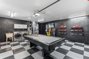Una mesa de billar en una habitación con piso a cuadros, una lavadora y estantes con coches de juguete.