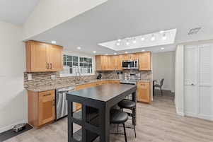 Cocina moderna con armarios de madera, una isla central, electrodomésticos de acero inoxidable y un comedor con mesa y sillas.