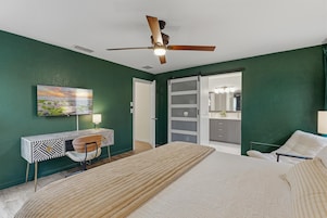 Un dormitorio con cama, escritorio, silla, ventilador de techo y puerta corredera que da a un baño.