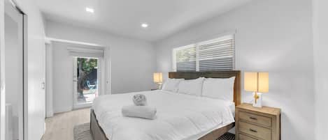 A bedroom with a bed, bedside tables, a window with blinds, and a door leading to an outdoor area.