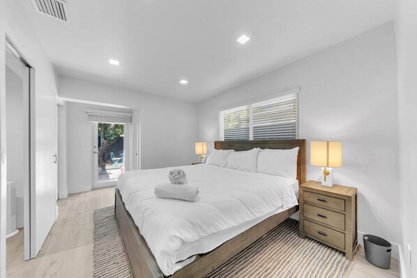A bedroom with a bed, bedside tables, a window with blinds, and a door leading to an outdoor area.