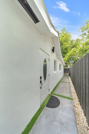 Un sendero conduce a un edificio blanco con una puerta y ventanas, una cerca de madera y un árbol al fondo.