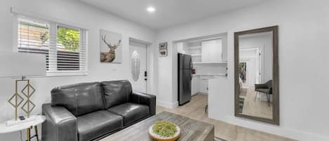 A modern living space with a leather sofa, a coffee table with a decorative bowl, a kitchen area with white cabinets, and a large mirror.