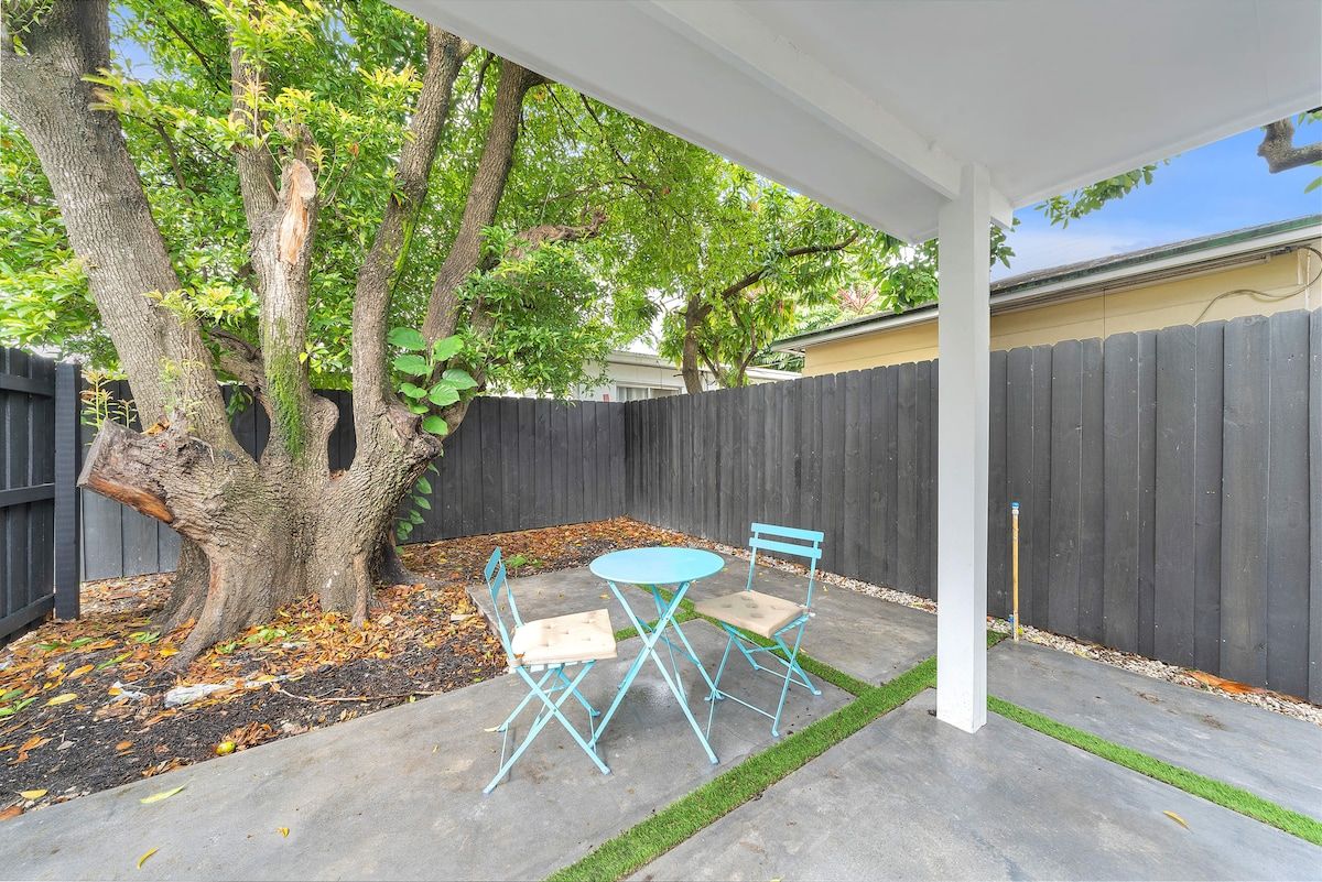 Un patio exterior con un árbol, una valla de madera y una pérgola blanca.
