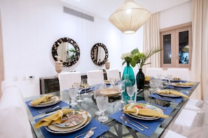 Comedor con mesa de vidrio dispuesta para una comida, dos espejos en la pared y un amplio ventanal con cortinas.