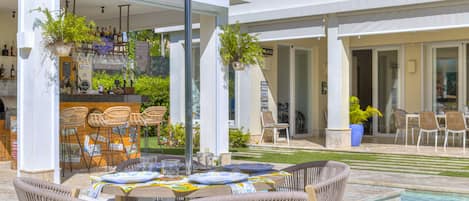 An outdoor dining area with wicker chairs and a table set for two, near a pool and a bar area with plants and bottles.