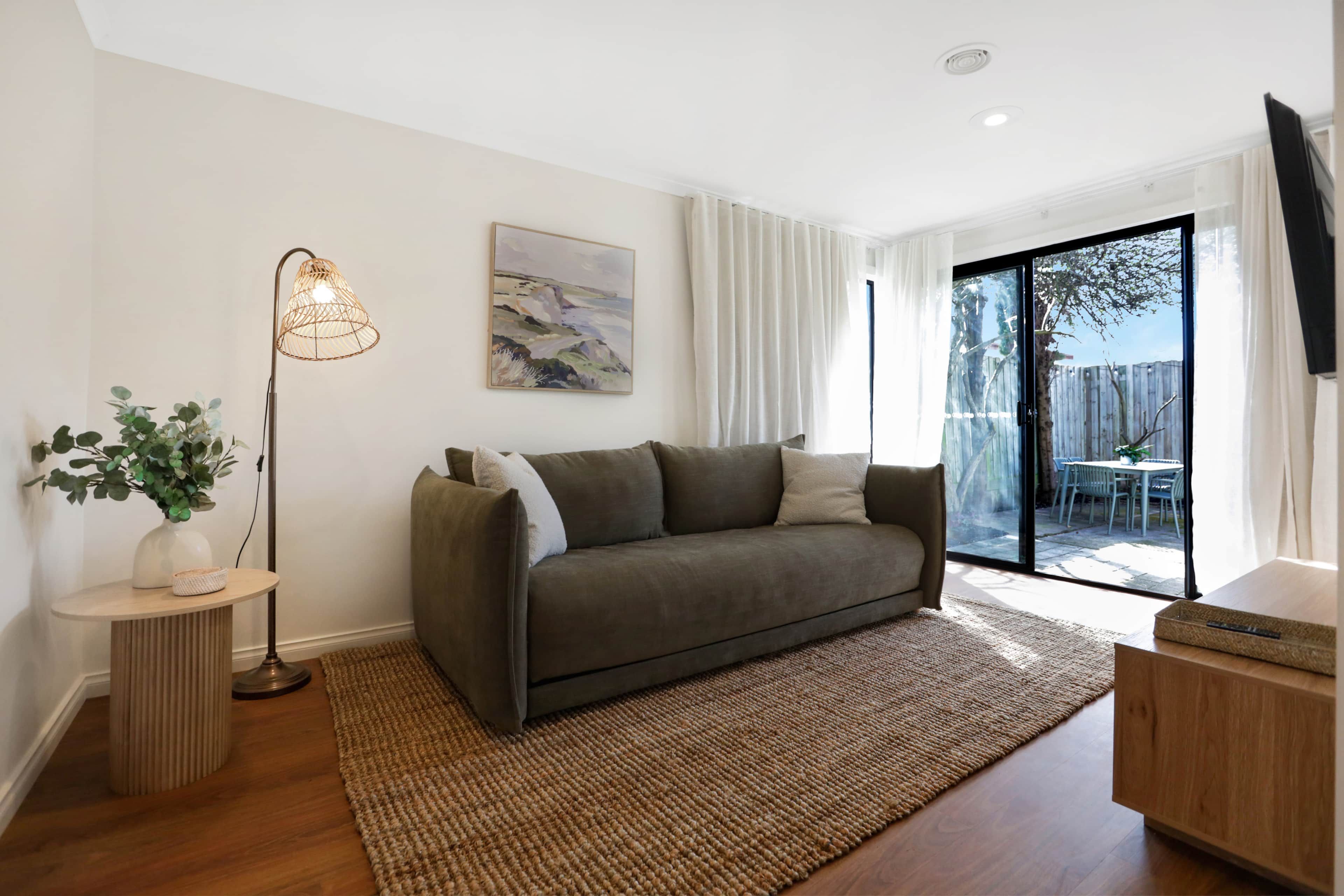 Ein modernes Wohnzimmer mit einer Couch, einer Stehlampe, einem Gemälde und Blick durch eine Glastür.
