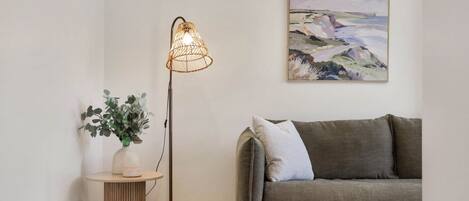 A room with a grey sofa, a woven floor lamp, a painting on the wall, a small round table with a vase and plant, and a wooden floor.