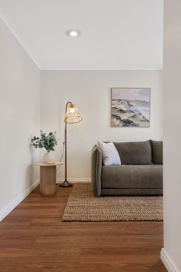 A room with a grey sofa, a woven floor lamp, a painting on the wall, a small round table with a vase and plant, and a wooden floor. - Fig Tree Cottage on Koroit | Central Warrnambool (Warrnambool)