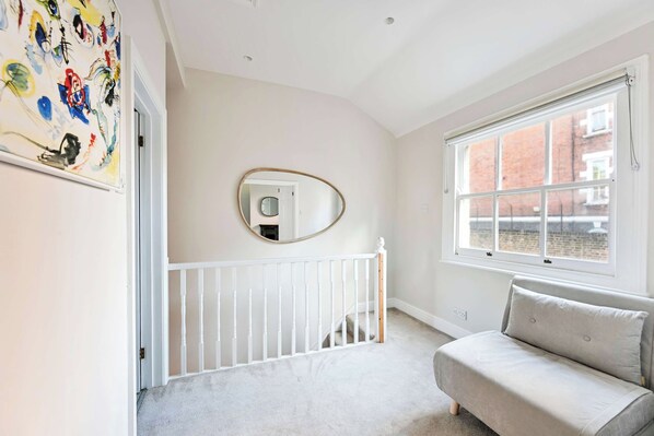 A room with a white crib, a round mirror on the wall, a window with a view of buildings, and a door with a colorful abstract painting. - Elegant St Johns Wood Mews-King Bed & Workspace (London)