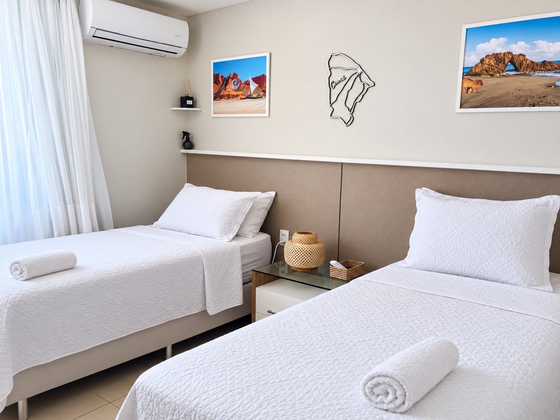 Quarto de hotel com duas camas, uma pequena mesa de cabeceira, ar condicionado fixado na parede e quadros emoldurados.