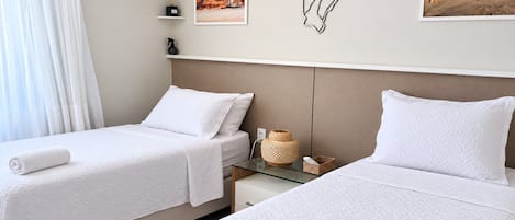 Quarto de hotel com duas camas, uma pequena mesa de cabeceira, ar condicionado fixado na parede e quadros emoldurados.