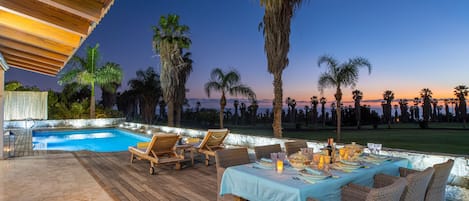 An outdoor dining area with a pool, wooden deck, and palm trees.