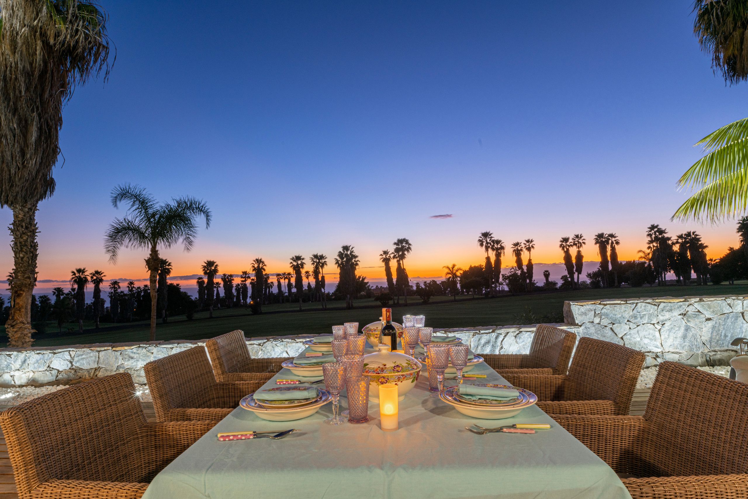 Un área de comedor al atardecer con muebles de mimbre, una mesa dispuesta con platos, vasos y una vela, con vista a un campo de golf.