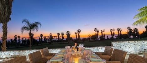 A sunset dining area with wicker furniture, a table set with plates, glasses, and a candle, overlooking a golf course.
