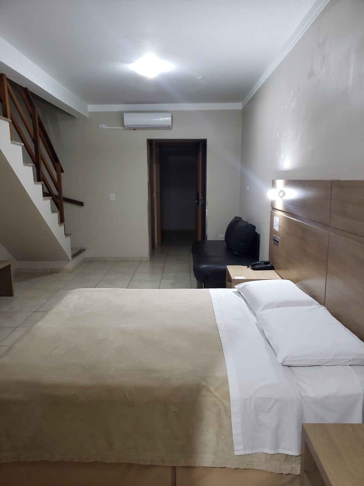 A hotel room with a bed, a desk, a chair, a wooden headboard, a staircase, and an air conditioning unit.