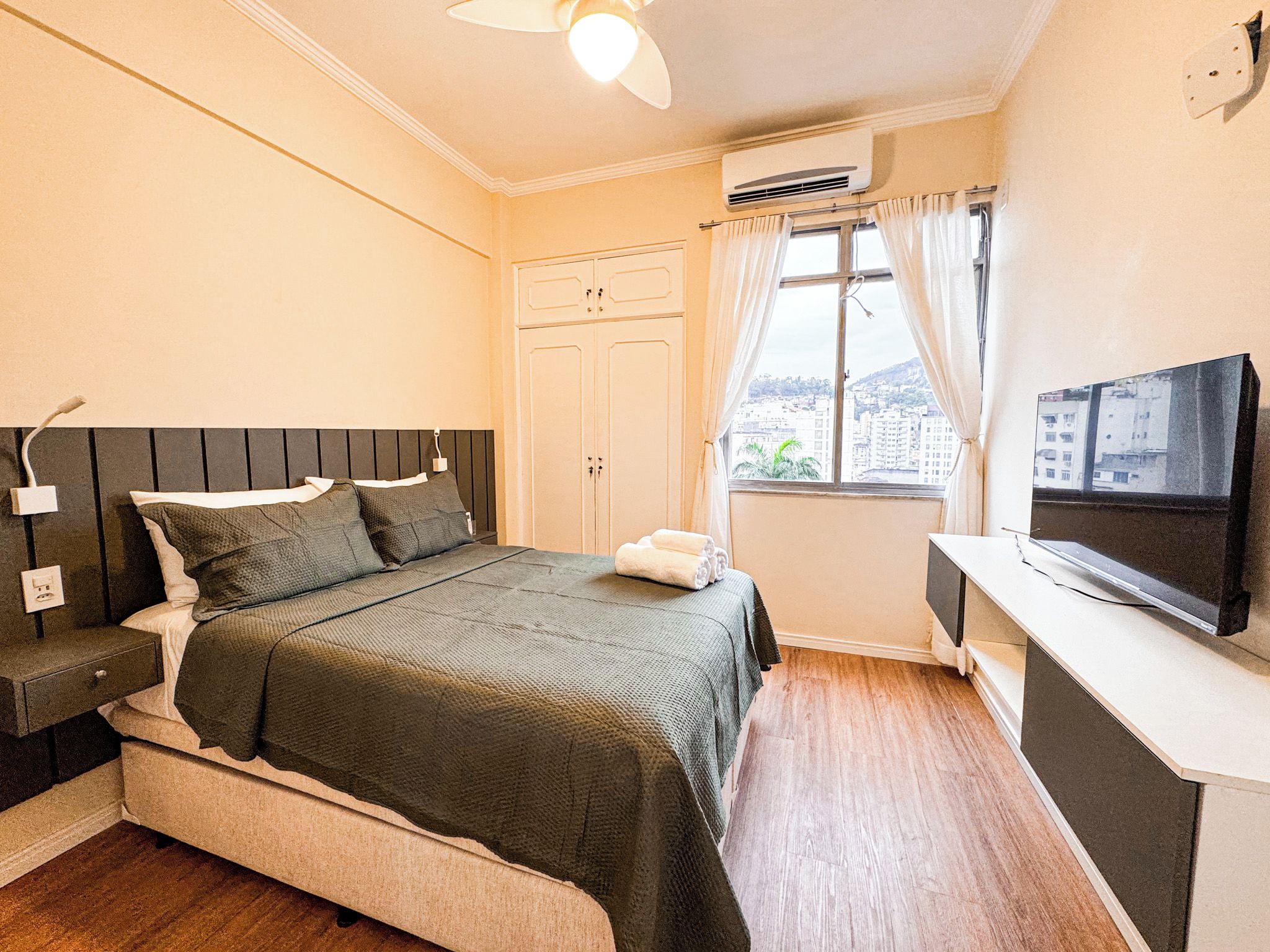 A bedroom with a bed, bedside table, TV, and a window with curtains.