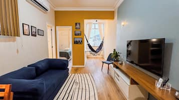 A modern living room with a sofa, a television, a hammock, and a view of the city through a window.