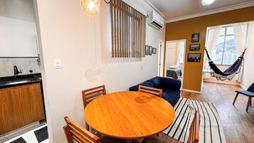 A dining area with a round wooden table and chairs, a kitchen with a sink and cabinets, a living area with a sofa and hammock, and a window with curtains.