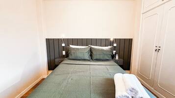 A neatly made bed with a green textured bedspread, two pillows, and a set of white towels on a blue mat.