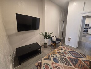 Ein Wohnzimmer mit einem an der Wand befestigten Flachbildfernseher, einer dunklen Unterhaltungseinrichtung, einer Topfpflanze, einem gemusterten Teppich und einer Tür, die in eine Küche führt.