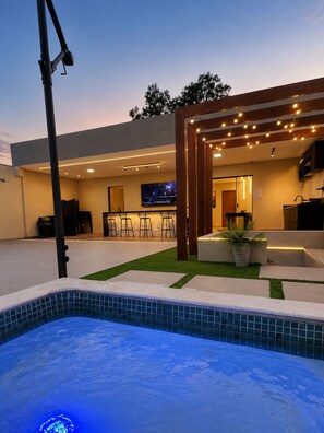 A modern outdoor pool area with a hot tub, bar area, and a television screen.