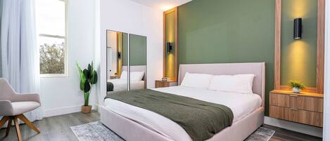 A modern bedroom with a large bed, a bedside table, a chair, a potted plant, and a mirror.