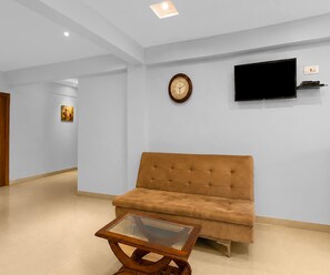A room with a sofa, a coffee table, a wall-mounted TV, and a clock. - Deluxe Room Hotel Naina Luxury Rooms (Hyderabad)