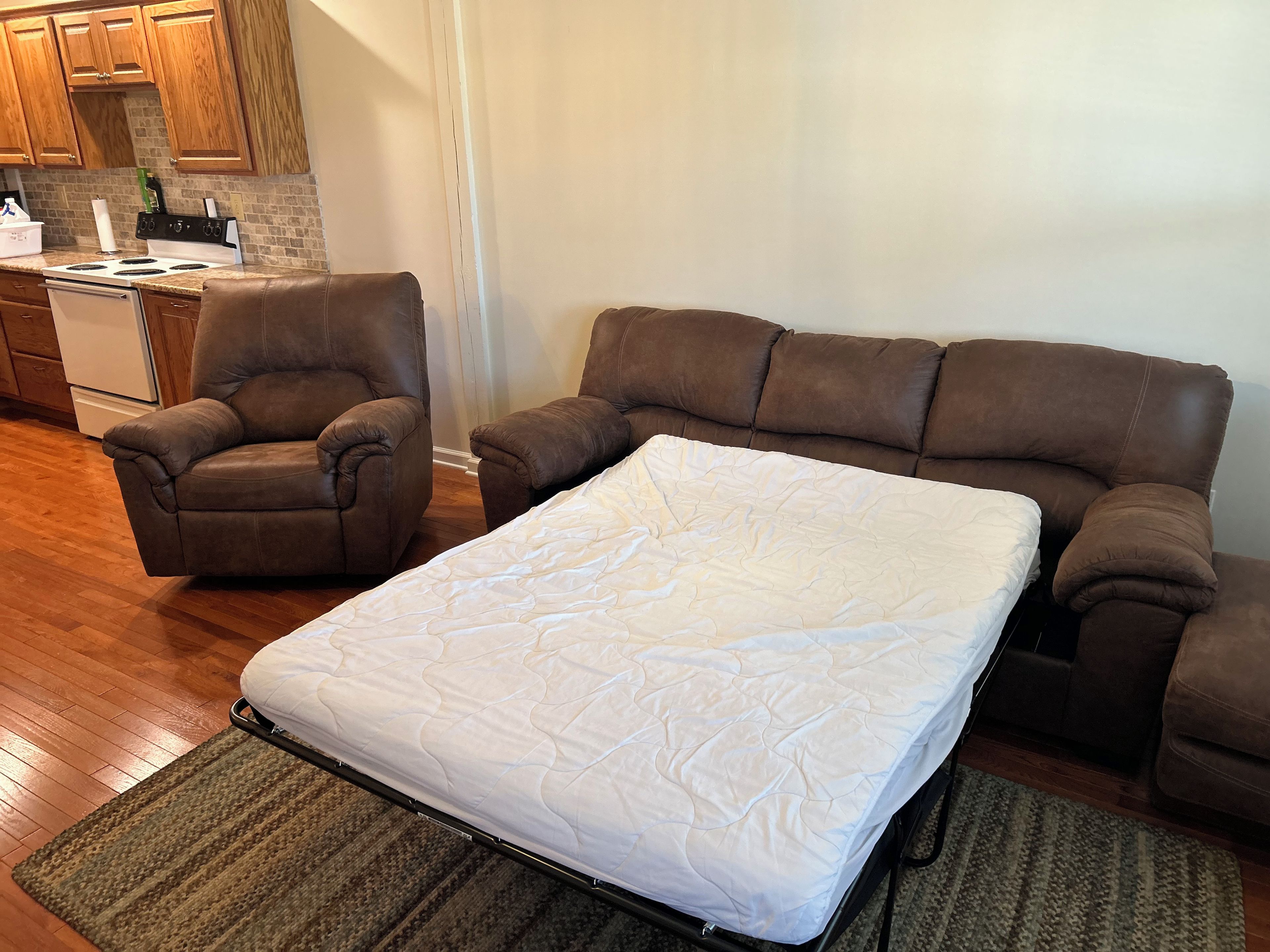 A living room with a sofa and a single armchair, a mattress on the floor, and a kitchen in the background.