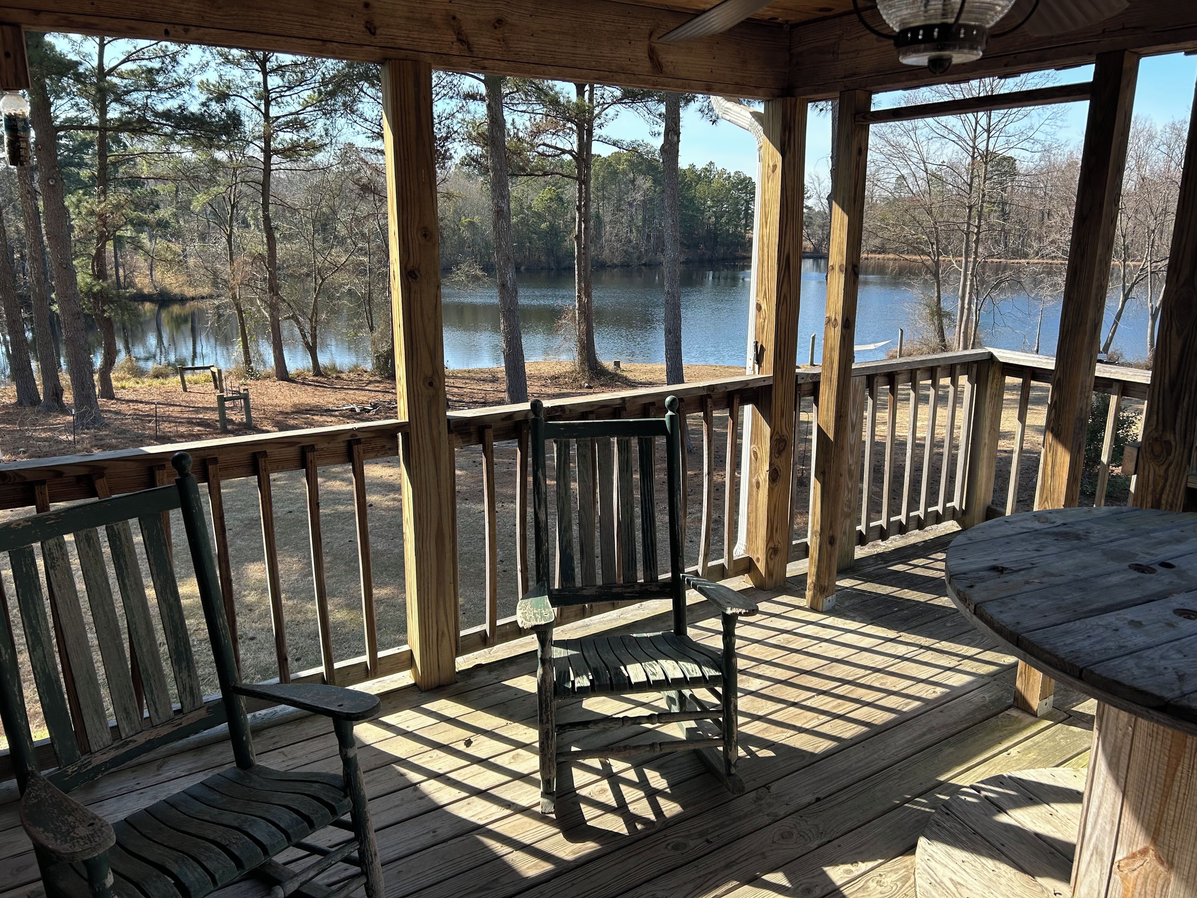 Terraza de madera con vista a un lago y árboles, un banco y una mesa.
