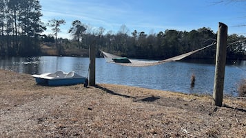Una hamaca atada entre dos árboles sobre un lago tranquilo, un bote amarrado en la orilla y una zona con césped y hojas secas.