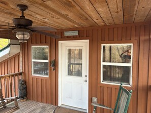 A wooden cabin with a white door, two windows with blinds, a ceiling fan, and a bear statue on the porch.