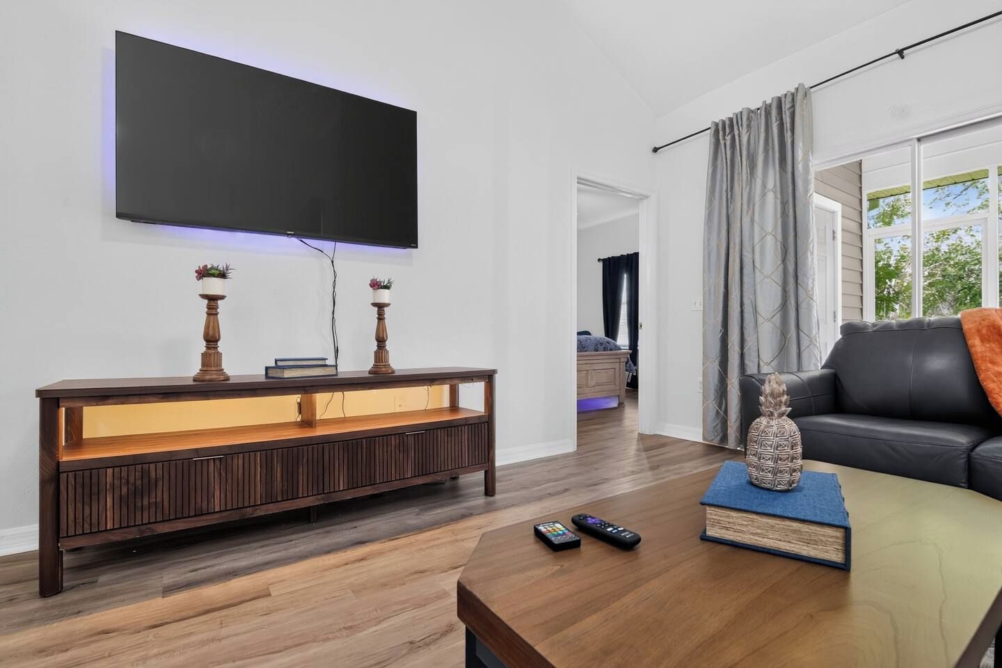 Ein modernes Wohnzimmer mit einem wandmontierten Flachbildfernseher, einem hölzernen Konsolentisch mit Dekorationselementen, einem Ledersofa und einem Couchtisch mit Fernbedienungen.