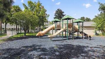 Ein Spielplatz mit Rutsche und Klettergerüsten, umgeben von Bäumen und einem Gebäude im Hintergrund.